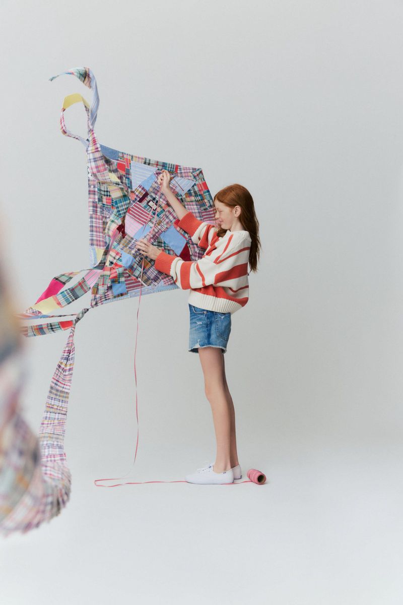 Girl with red hair in a striped sweater holding a colorful patchwork kite against a white seamless studio background.