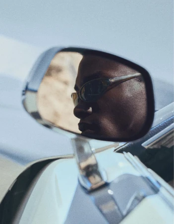 Reflection of a Black man wearing wraparound sunglasses seen in the chrome side mirror of a car.
