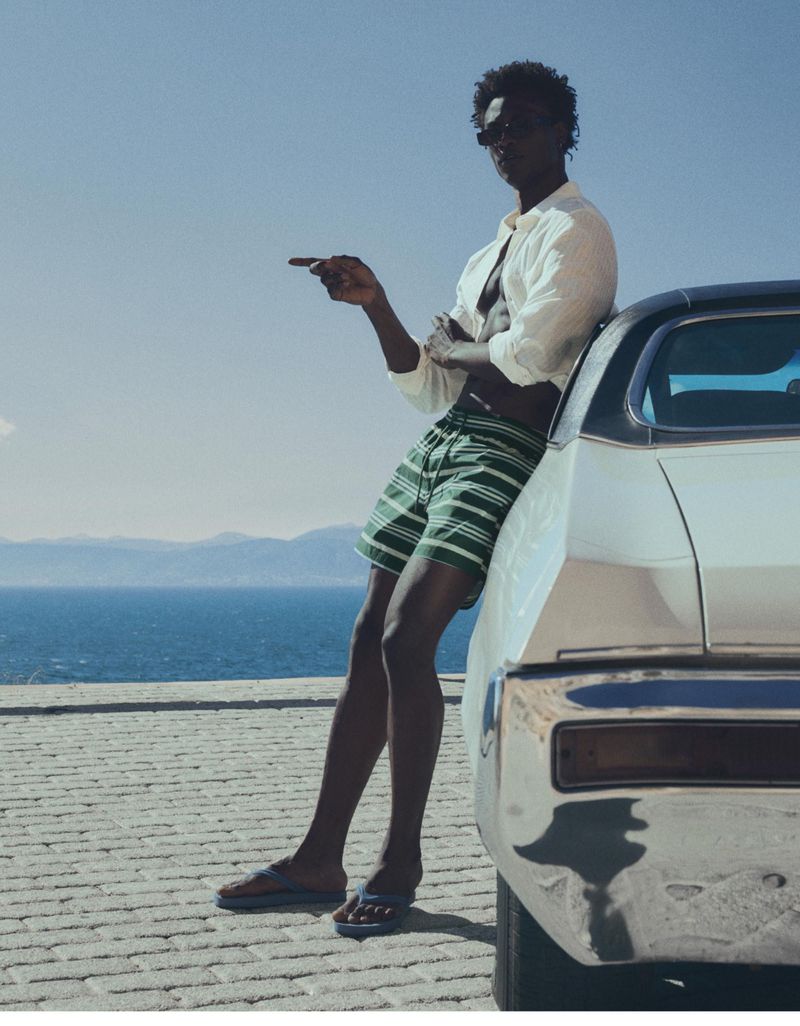 Model George Godspower wearing striped swim shorts and an open white shirt, leaning on a classic white car overlooking the ocean.