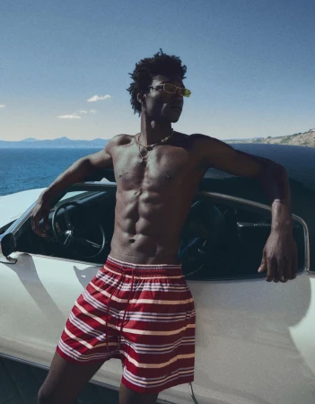 Model George Godspower in striped swim shorts and yellow sunglasses leaning against a white car by the ocean.