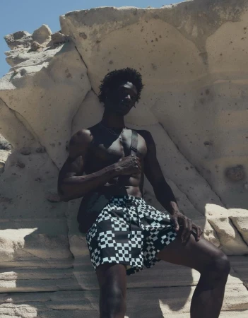 Model George Godspower wearing black and white checkered swim shorts against pale coastal rock formations in bright sun.