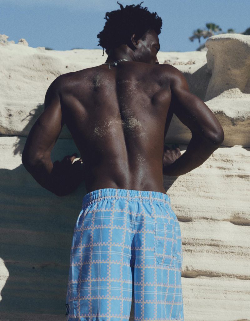 Shirtless Black male model seen from behind wearing light blue plaid swim trunks while leaning against pale coastal rock.