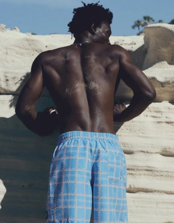 Shirtless Black male model seen from behind wearing light blue plaid swim trunks while leaning against pale coastal rock.