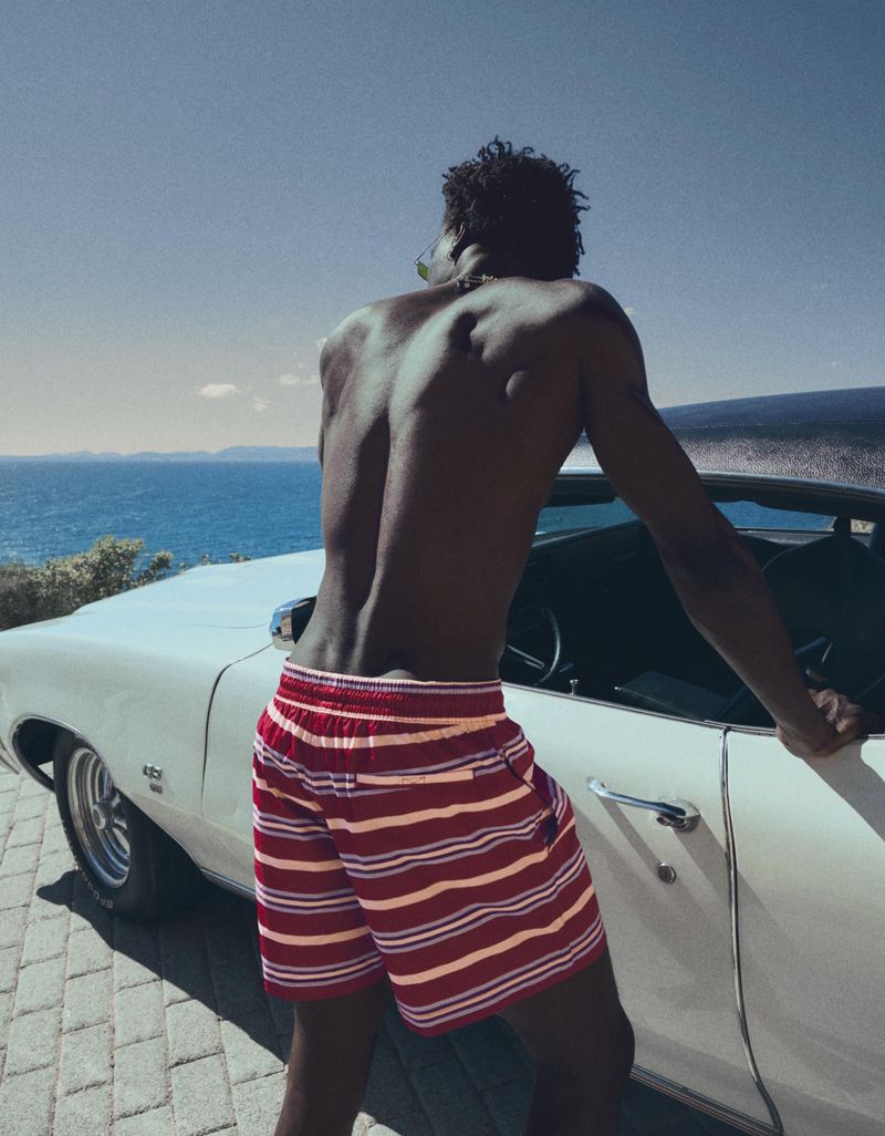 Shirtless Black man in red striped swim shorts leaning against a vintage white convertible car overlooking the blue ocean.