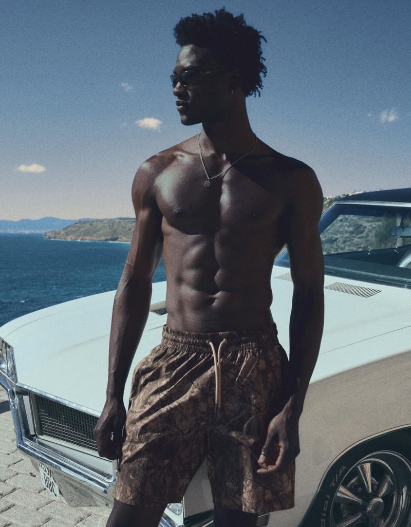Shirtless man in patterned brown swim shorts and sunglasses posing in front of a vintage classic car by the coast