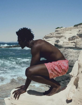 A Black man wearing pink patterned swim shorts crouched on a pale rocky cliff beside the ocean.