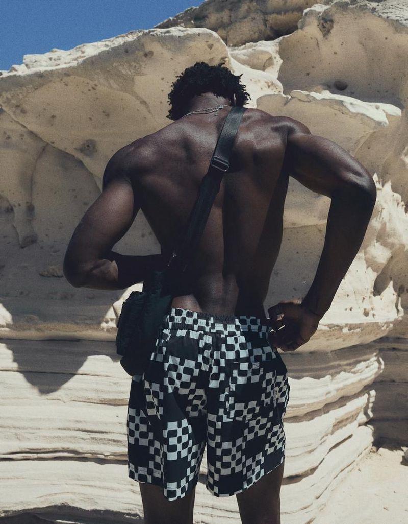 Model George Godspower seen from behind wearing black and white checkered swim shorts against pale coastal rock formations.