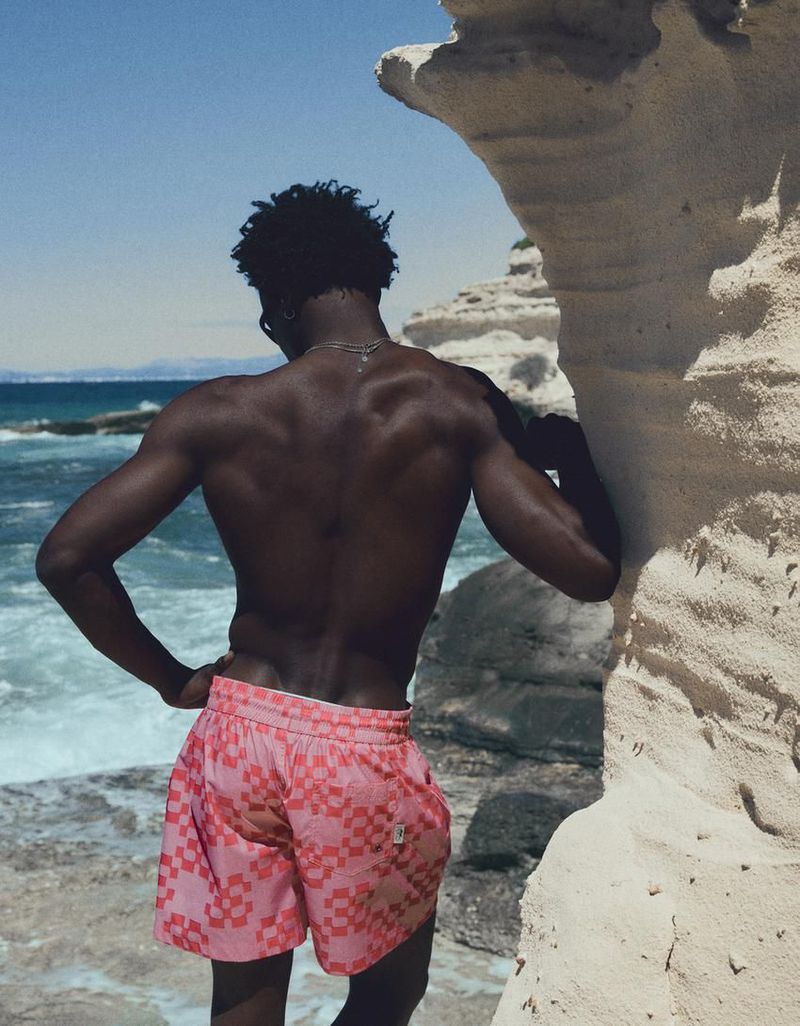 Model George Godspower from behind in pink patterned swim shorts standing against a sandstone coastal rock formation near the ocean.