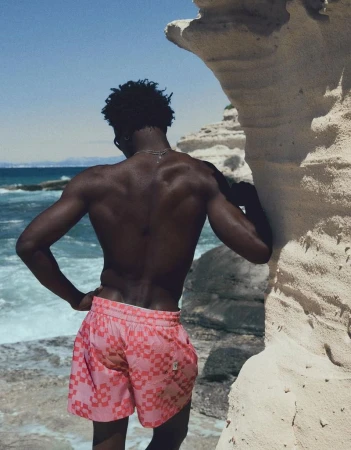 Model George Godspower from behind in pink patterned swim shorts standing against a sandstone coastal rock formation near the ocean.