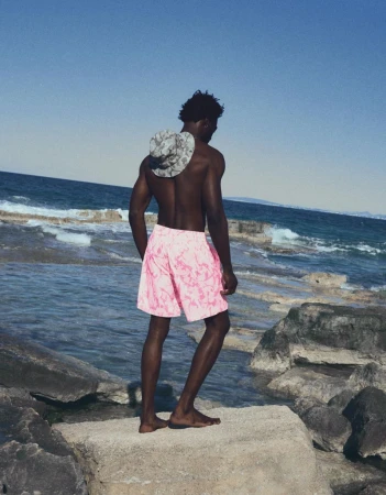 Model George Godspower seen from behind in pink swim shorts and a bucket hat standing on coastal rocks overlooking the sea.