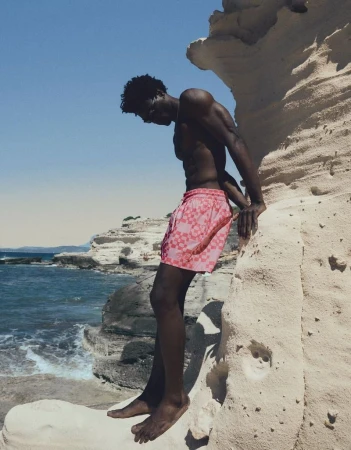 Model George Godspower in pink patterned swim shorts standing barefoot against a white coastal cliff beside the ocean.