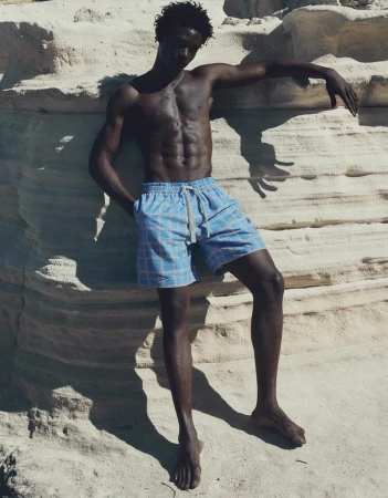 Model George Godspower wearing patterned light blue swim shorts leaning against a textured pale rock cliff.