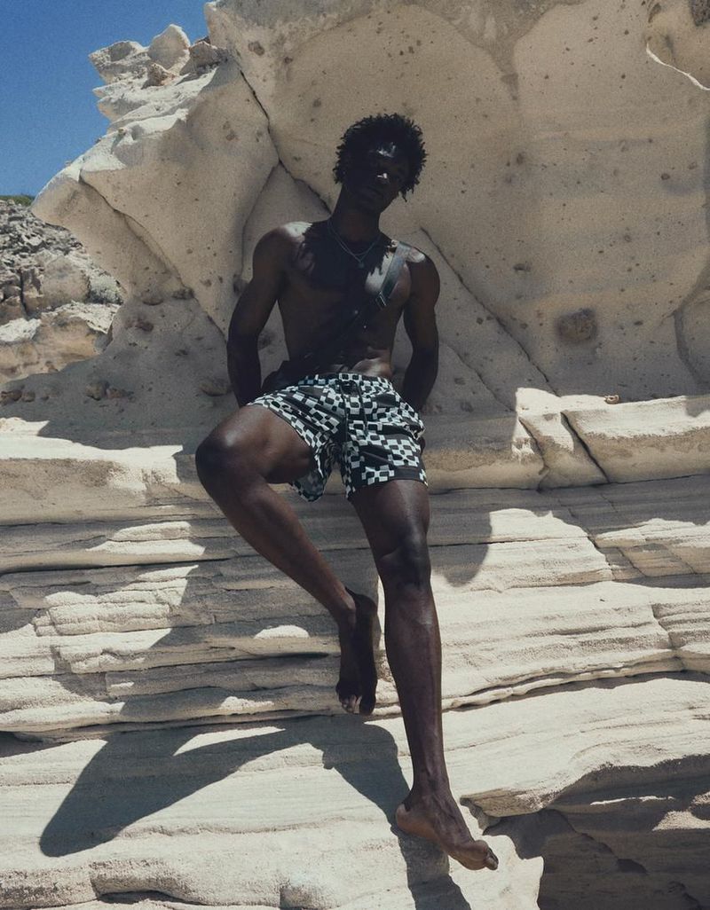 Black man wearing checkered swim shorts posing against weathered white rock formations under bright sun