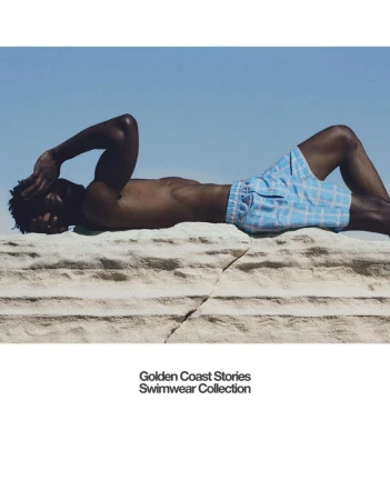Shirtless Black man in light blue patterned swim shorts reclining on a textured white rock under a clear blue sky.