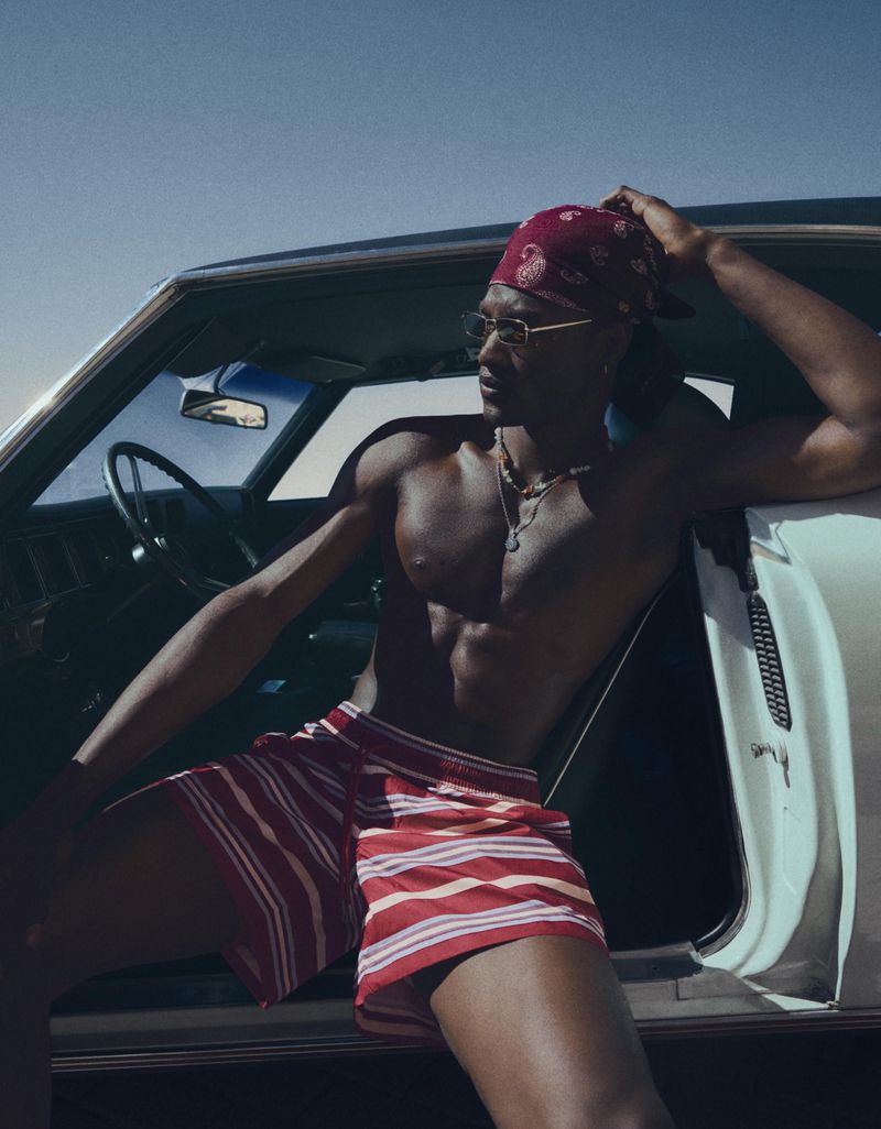 Shirtless Black man wearing a red bandana and striped swim shorts seated in the open doorway of a classic car under a clear sky.