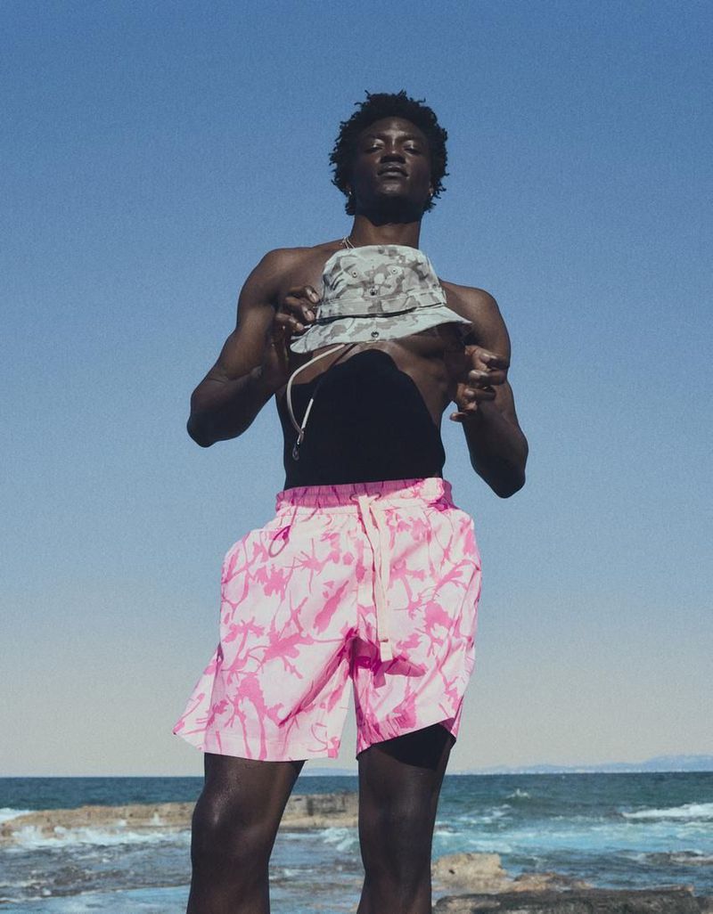 Model George Godspower in pink patterned swim shorts holding a bucket hat against an ocean backdrop.