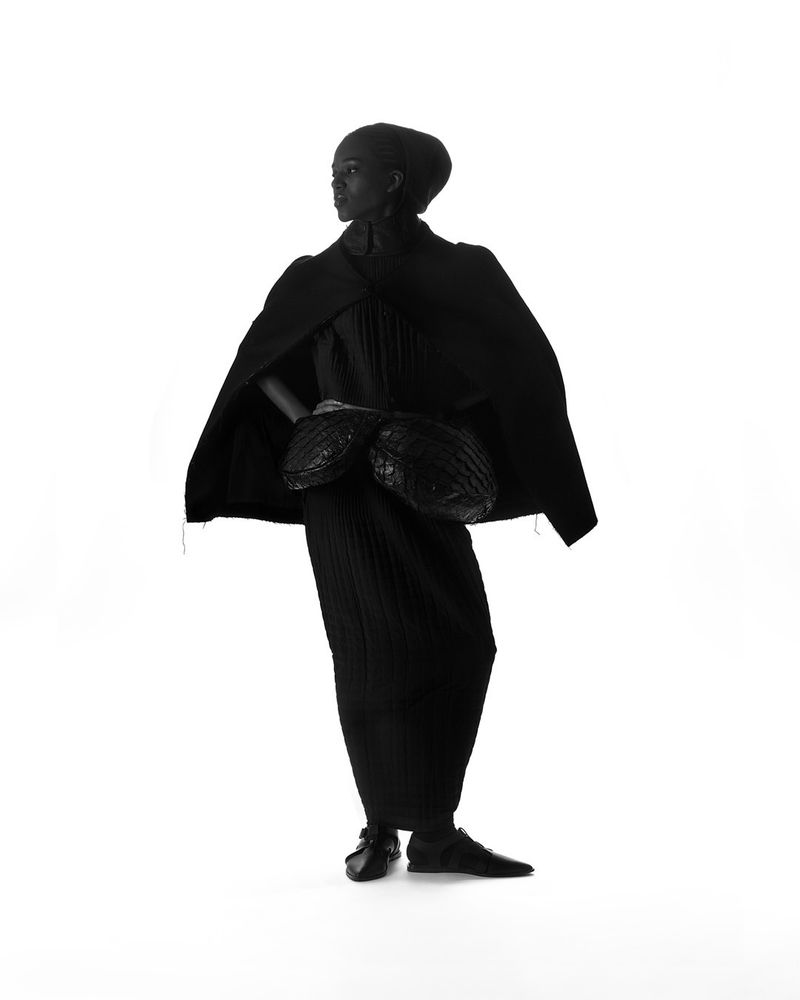 Model wearing an all-black draped ensemble with a cape and waist bag against a white seamless background.