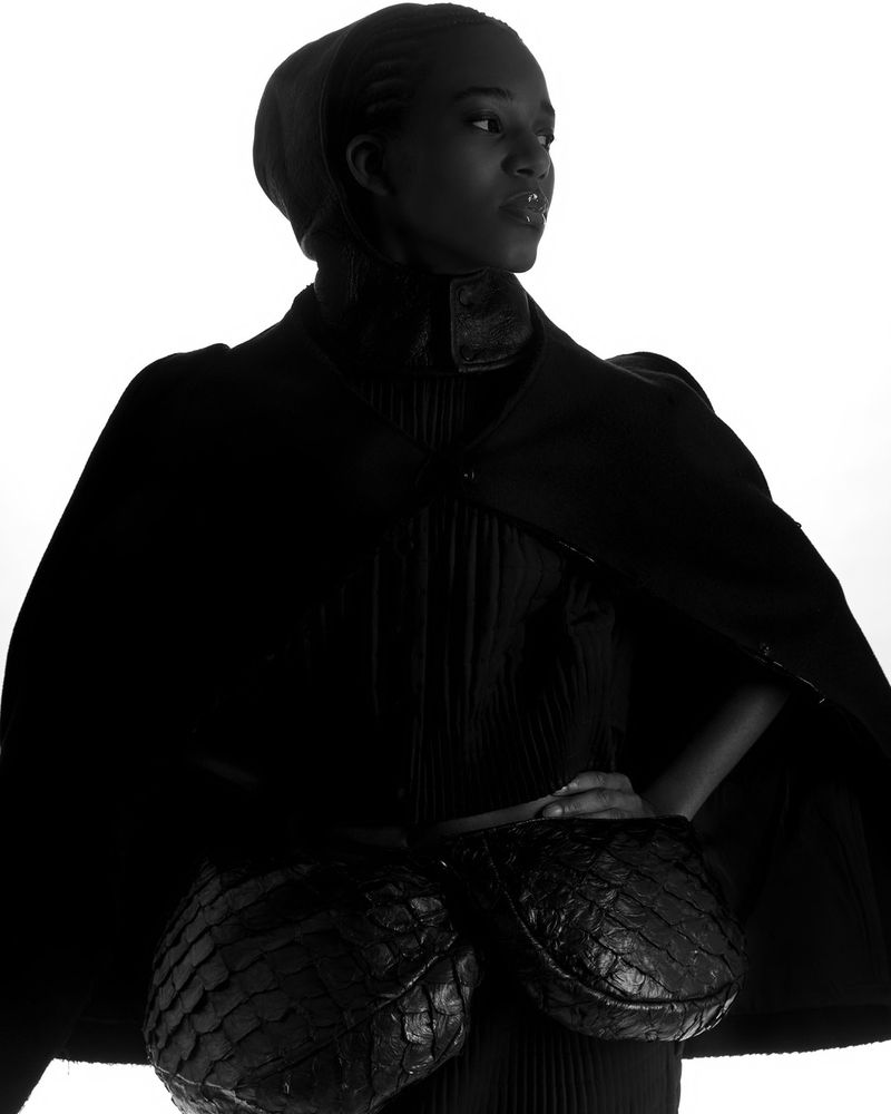 Model Sara Felipe wearing a structured black cape and textured waist pockets against a white backdrop for Osklen lookbook.