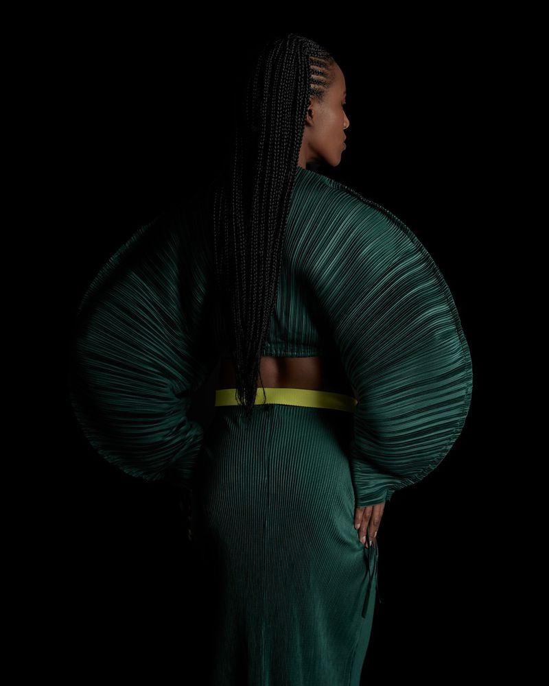 Model Sara Felipe seen from behind wearing an emerald green pleated garment with dramatic sculptural sleeves against a black backdrop.
