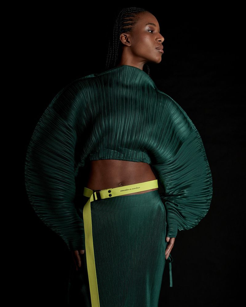 Model Sara Felipe wears a sculptural pleated dark green cropped top and matching skirt with a neon yellow belt against a black backdrop.