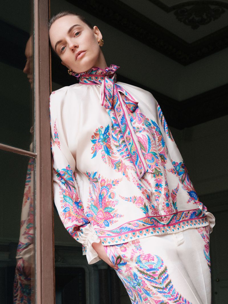 Model Fran Summers wearing a cream paisley silk blouse with a matching tied scarf collar leaning against a door frame.