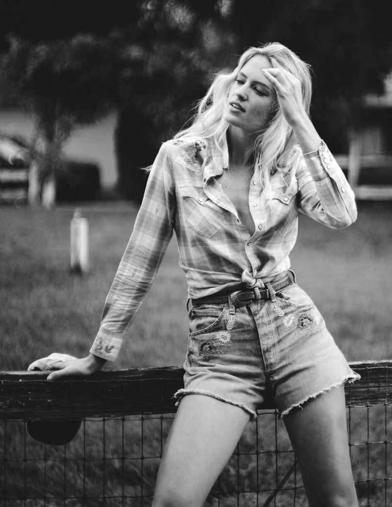 Model Kat Carter wearing a tied plaid shirt and embroidered denim cutoffs while leaning against a wooden fence.
