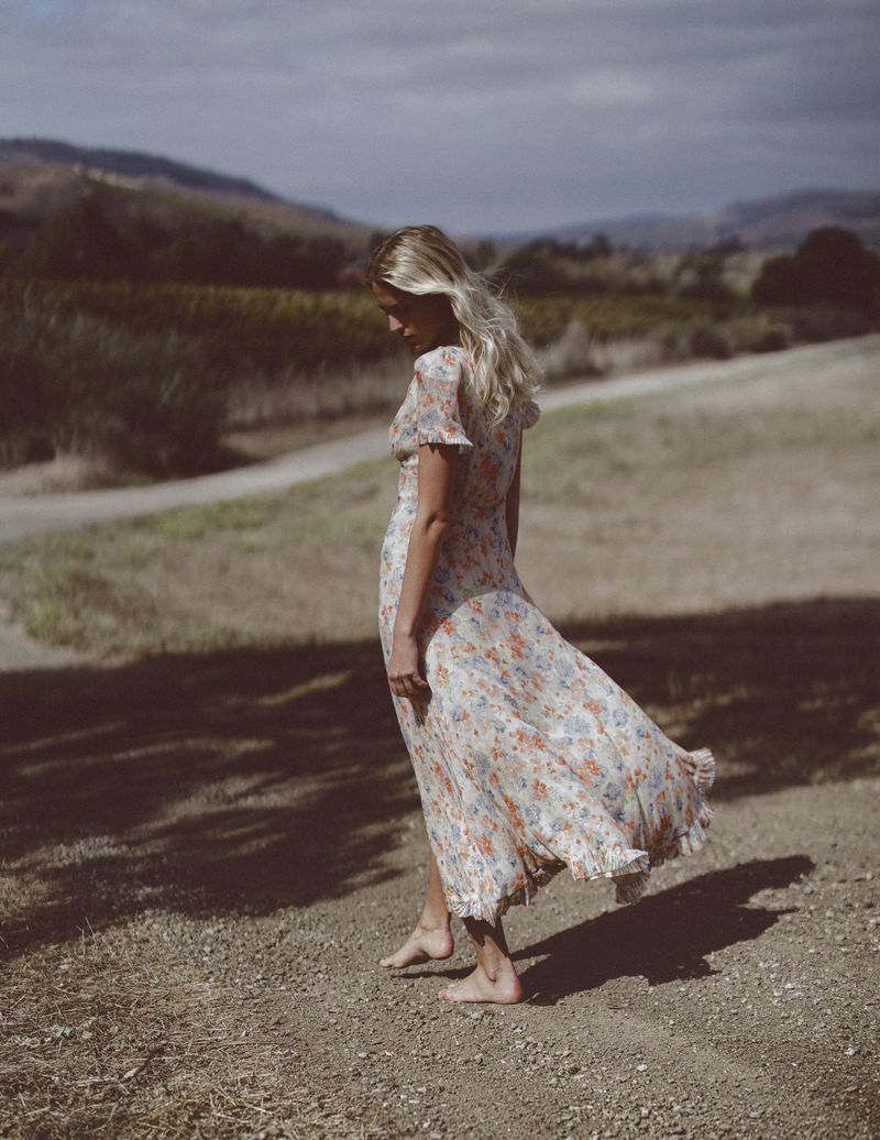 Model Kat Carter walking barefoot along a rural path wearing a flowing floral dress in front of distant hills.