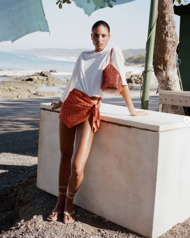 Michiko Foy wearing a white t-shirt and rust wrap skirt leaning on a white block near a coastal beach for Christopher Esber.