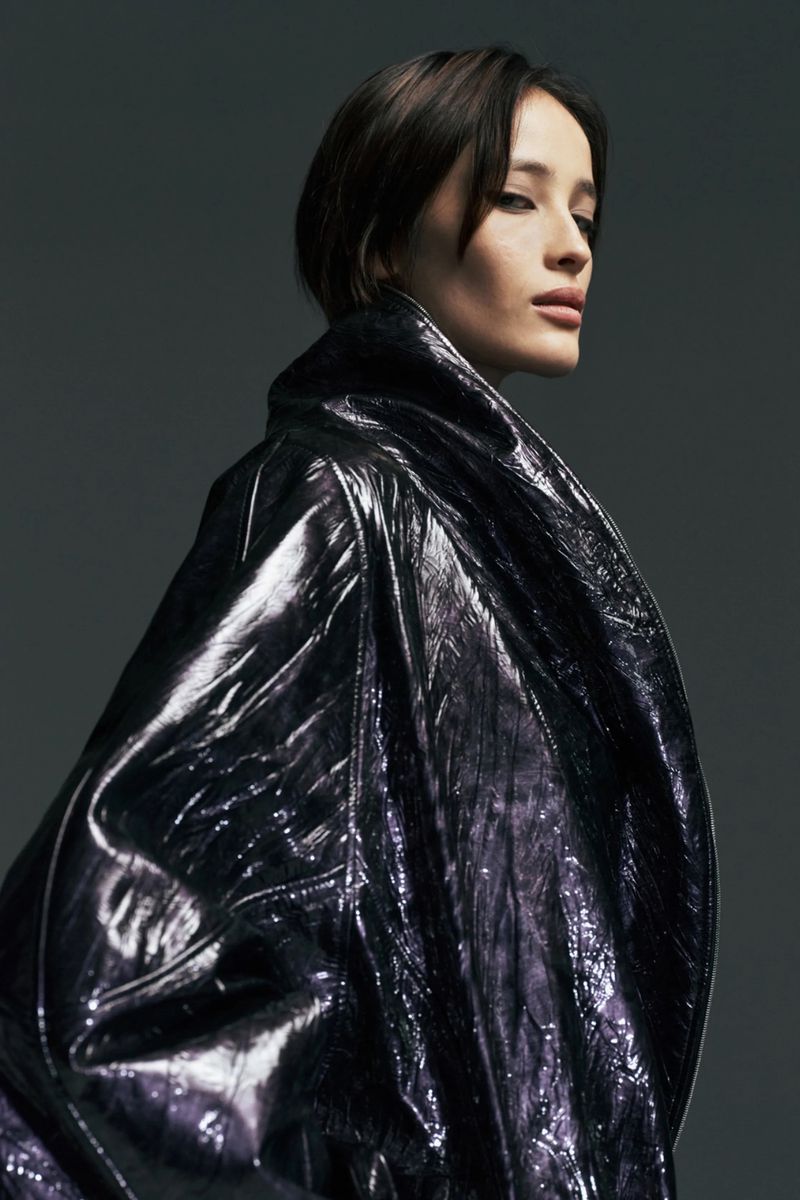 Portrait of a female model in a glossy, dark trench coat in a studio with a grey background, looking over her shoulder.