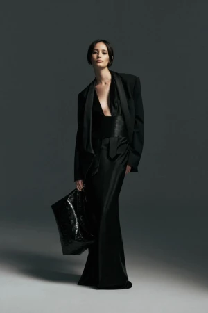Model wearing a tailored black blazer and floor-length black satin gown holding a glossy black handbag against a grey backdrop.