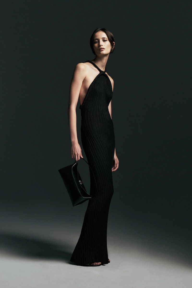 Model poses in a floor-length black ribbed halter dress holding a black leather handbag against a dark studio background.