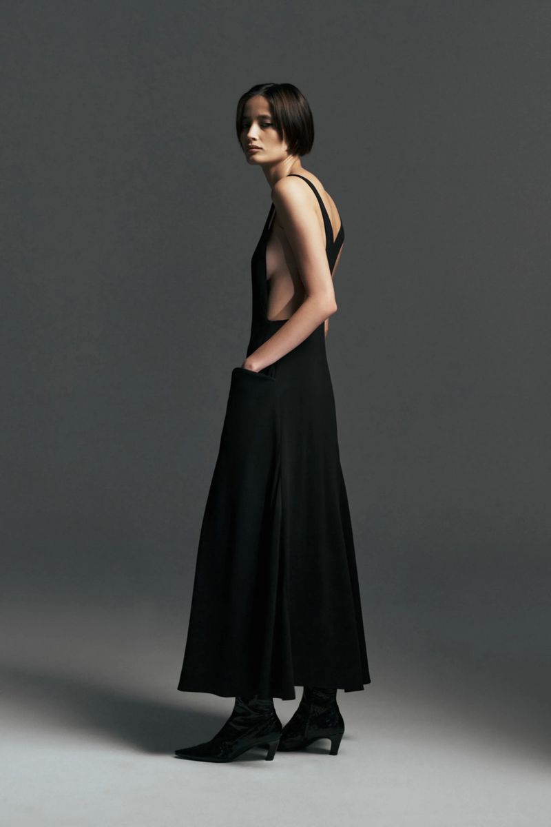 Model wearing a sleeveless black open-back maxi dress with pockets and black pointed-toe boots against a grey backdrop.