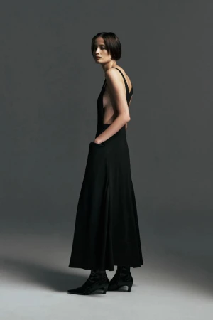 Model wearing a sleeveless black open-back maxi dress with pockets and black pointed-toe boots against a grey backdrop.