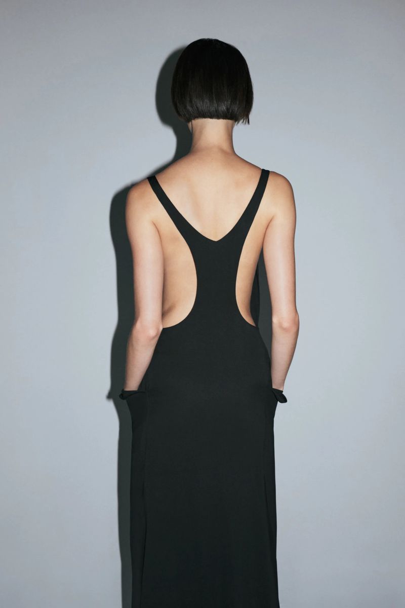Model with a short dark bob seen from the back wearing a black backless cutout dress against a grey studio backdrop.