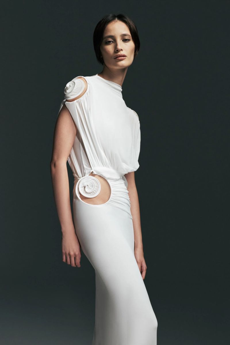Model wearing a draped white Christopher Esber gown with a circular waist cutout and rosette accents against a dark studio background.
