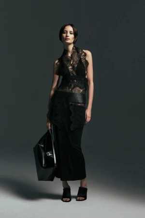 Model wearing a black sheer lace top, long black skirt, and open-toe heels while holding a large black tote against a grey backdrop.
