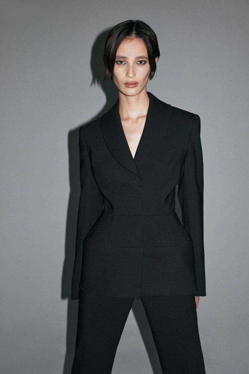 A model poses wearing a structured black tailored blazer suit against a neutral grey studio backdrop with directional shadows.