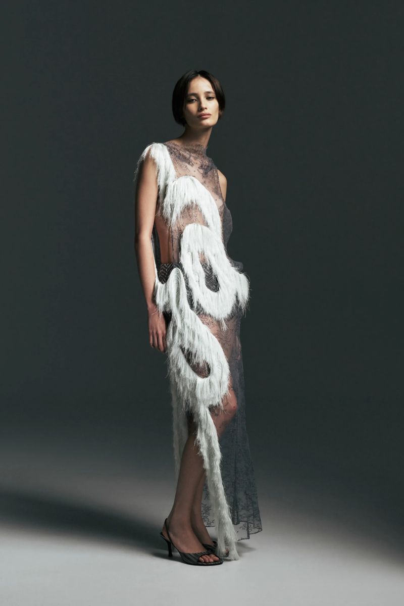 Model wearing a sheer lace dress with white curved fringe detailing and black strappy heels against a dark grey studio background.