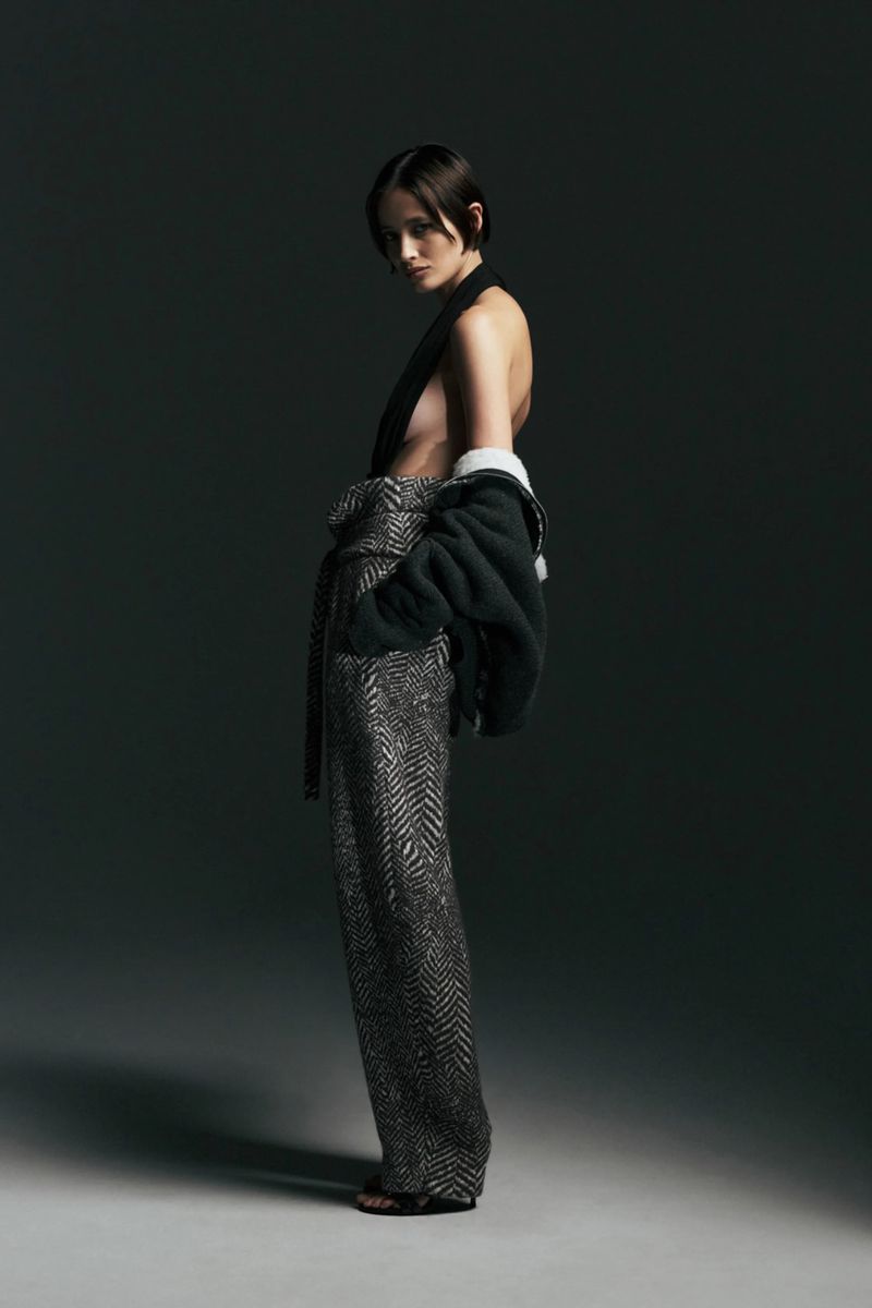 Model wearing high-waisted patterned wide-leg trousers and a dark jacket off the shoulder against a studio backdrop.