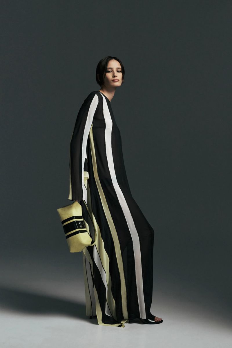 Model in a black, white, and pale yellow striped floor-length dress holding a matching clutch bag against a grey studio backdrop.