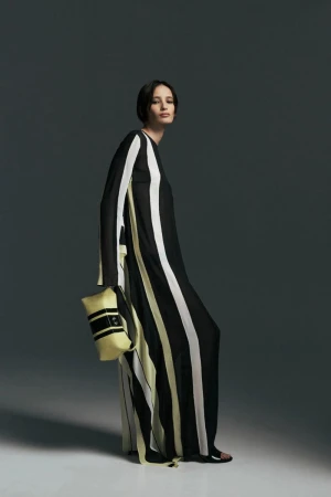 Model in a black, white, and pale yellow striped floor-length dress holding a matching clutch bag against a grey studio backdrop.