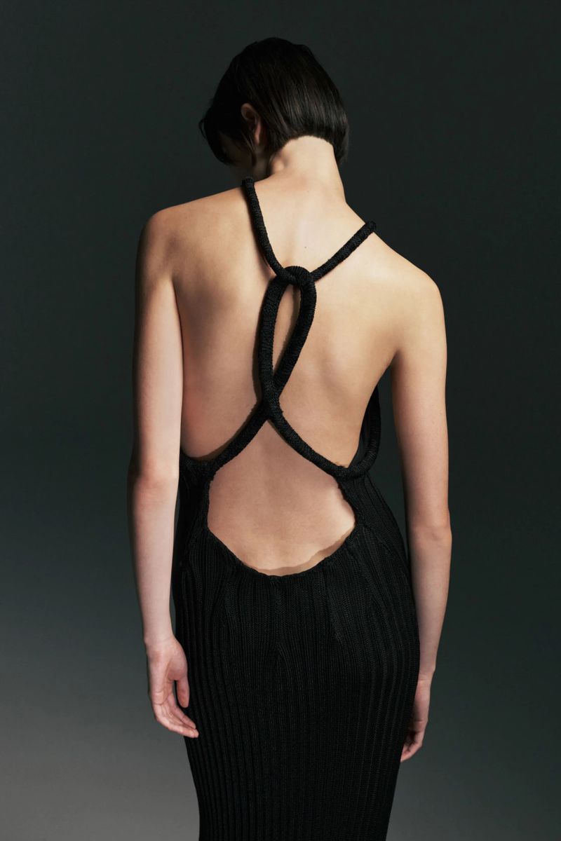 Rear view of a model with short dark hair wearing a backless black ribbed knit dress with knotted back straps against a dark studio backdrop.