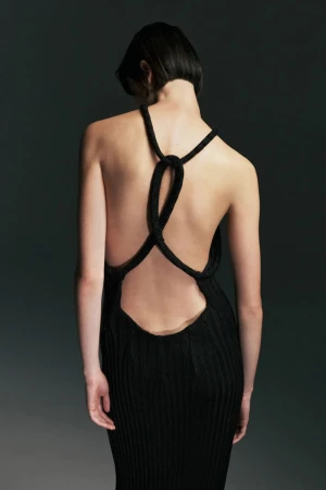 Rear view of a model with short dark hair wearing a backless black ribbed knit dress with knotted back straps against a dark studio backdrop.