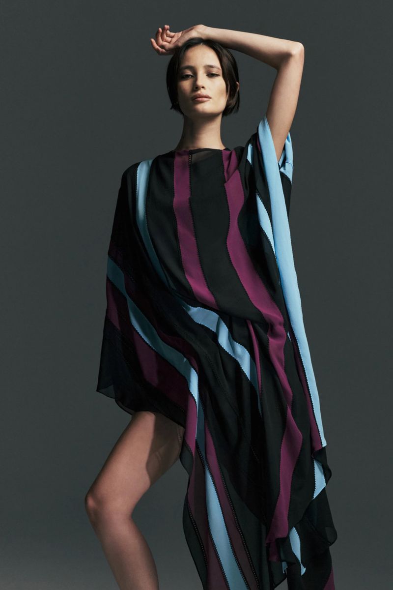 Model wearing an asymmetric draped striped dress in black, plum, and light blue against a dark grey studio background.