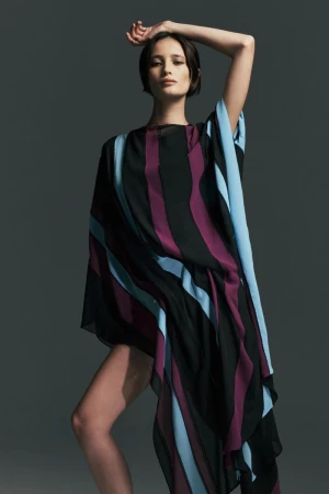Model wearing an asymmetric draped striped dress in black, plum, and light blue against a dark grey studio background.