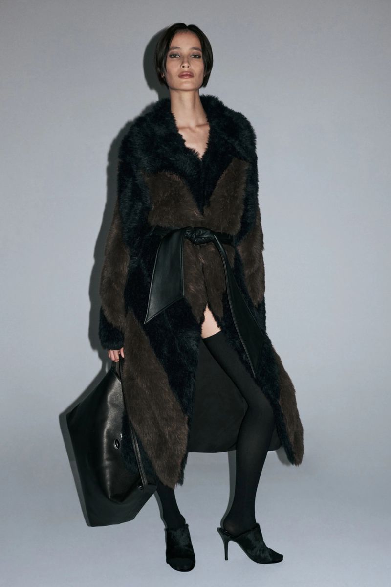 Model wearing a belted two-tone faux-fur coat, black tights, and heels while holding a large black bag against a grey background.