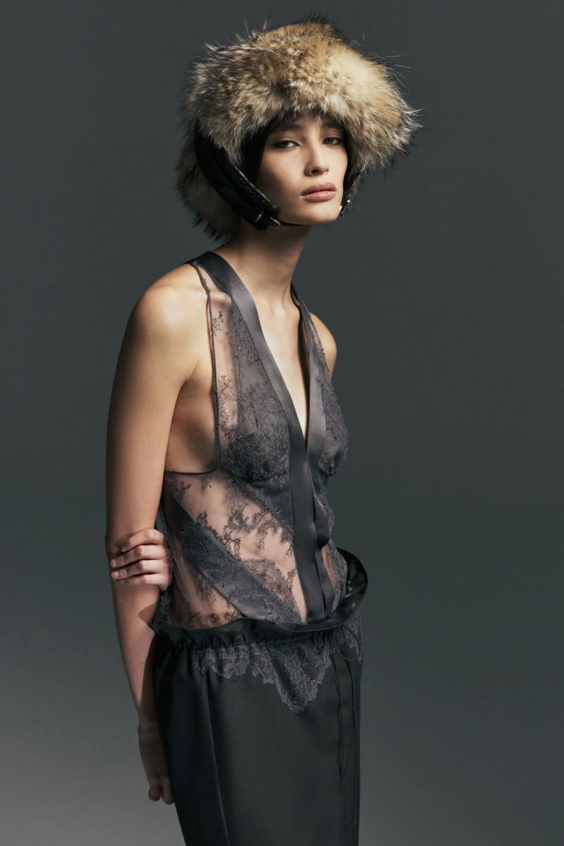 Model wearing a sheer grey lace top and an oversized fur trapper hat against a dark grey studio background.