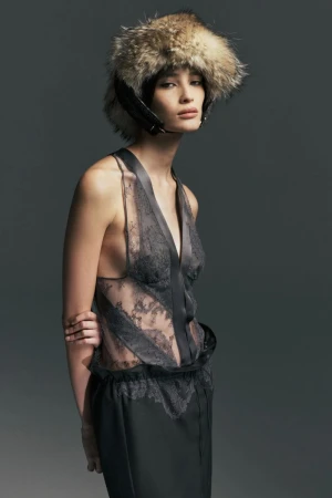 Model wearing a sheer grey lace top and an oversized fur trapper hat against a dark grey studio background.