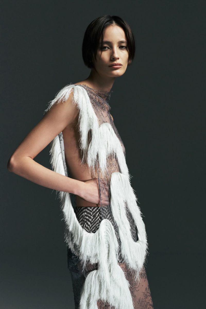 Model posing in a sheer lace top accented with looped white fringe against a dark grey studio background.