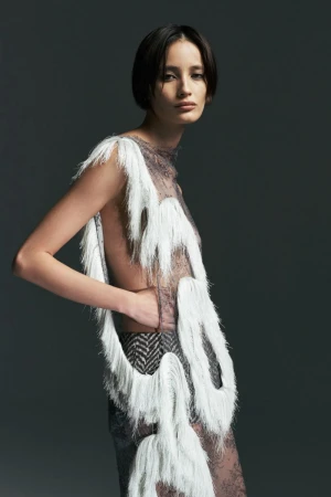 Model posing in a sheer lace top accented with looped white fringe against a dark grey studio background.